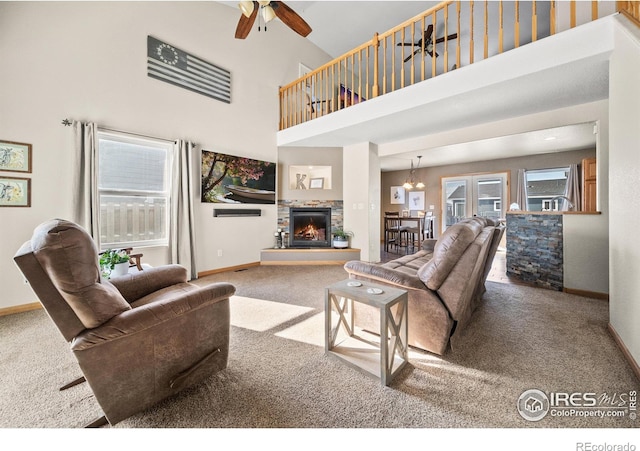 carpeted living room featuring ceiling fan with notable chandelier, plenty of natural light, a stone fireplace, and a towering ceiling