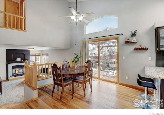 dining room with a high ceiling, a ceiling fan, baseboards, light wood-style floors, and a glass covered fireplace