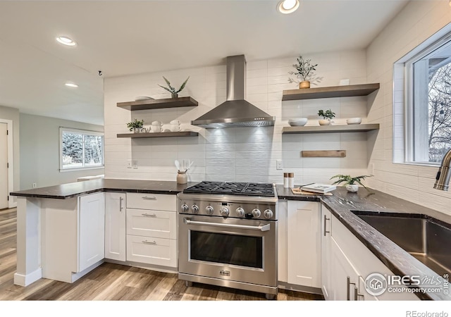 kitchen featuring stainless steel stove, white cabinetry, wall chimney range hood, sink, and backsplash