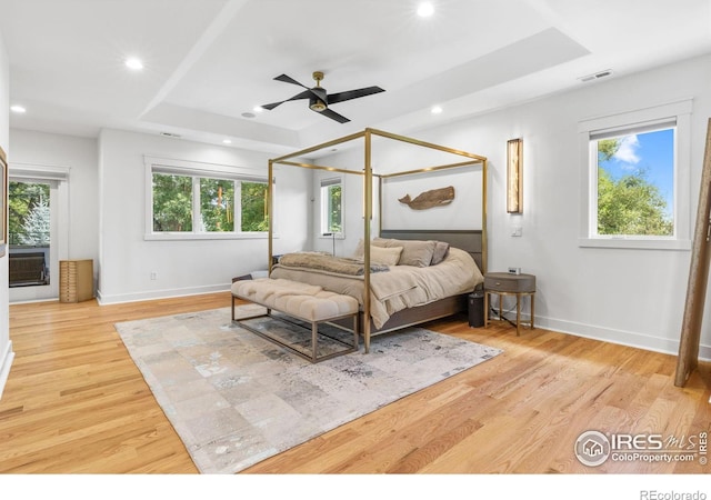 bedroom with ceiling fan, a raised ceiling, light hardwood / wood-style flooring, and multiple windows