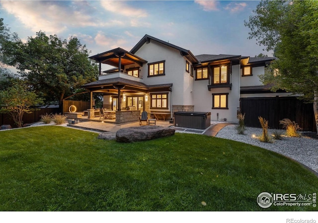 back house at dusk with a balcony, a patio area, a lawn, and a hot tub