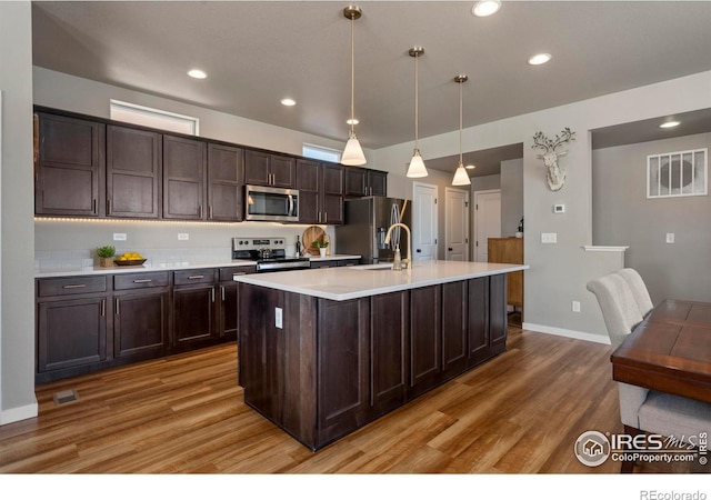 kitchen with decorative light fixtures, dark brown cabinets, a center island with sink, stainless steel appliances, and hardwood / wood-style floors