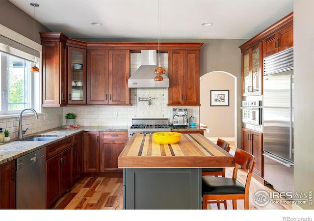 kitchen with appliances with stainless steel finishes, sink, a center island, light stone countertops, and wall chimney range hood