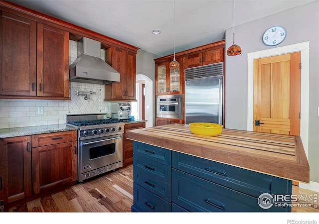 kitchen with wall chimney range hood, light hardwood / wood-style flooring, hanging light fixtures, high end appliances, and tasteful backsplash