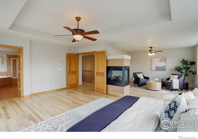 bedroom with a multi sided fireplace, ensuite bathroom, ceiling fan, a raised ceiling, and light wood-type flooring