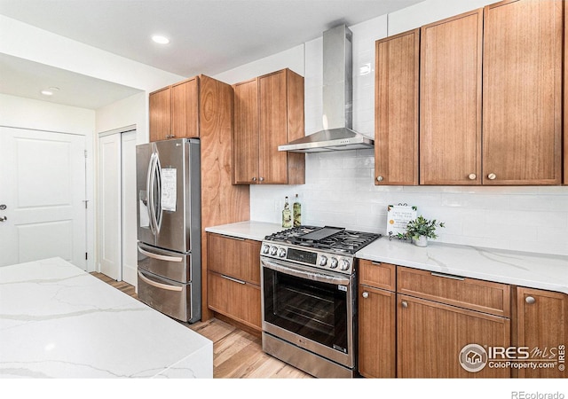 kitchen with appliances with stainless steel finishes, light wood-type flooring, wall chimney range hood, light stone countertops, and decorative backsplash