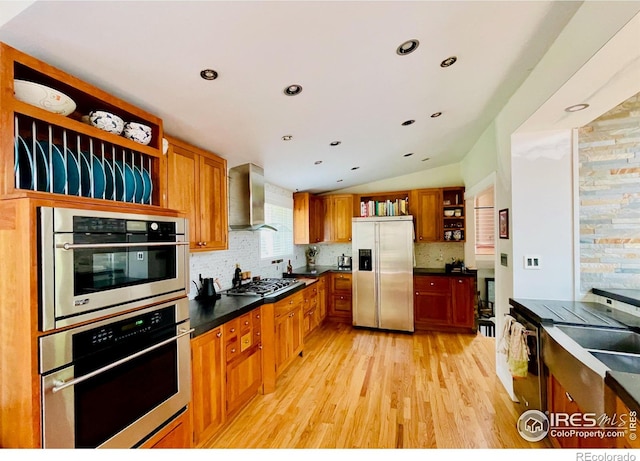 kitchen featuring lofted ceiling, light wood-type flooring, appliances with stainless steel finishes, decorative backsplash, and wall chimney range hood