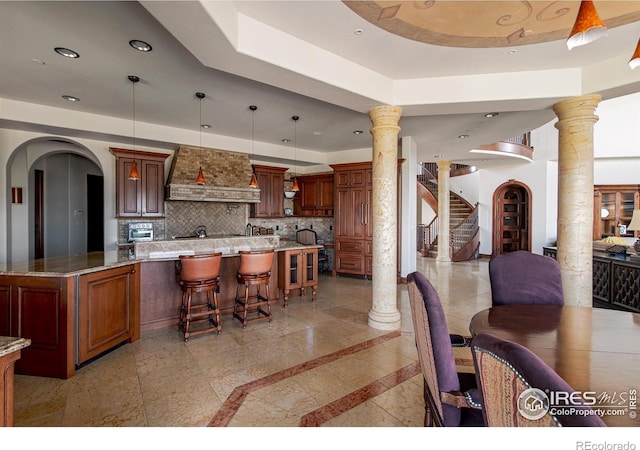 kitchen featuring ornate columns, premium range hood, pendant lighting, tasteful backsplash, and a breakfast bar area