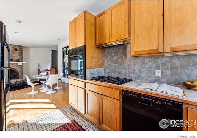 kitchen with a brick fireplace, backsplash, black appliances, and light wood-type flooring