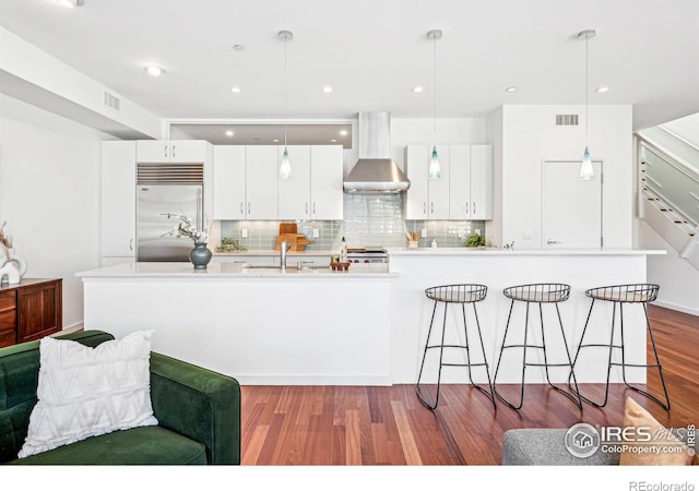 kitchen featuring built in fridge, wall chimney range hood, pendant lighting, and white cabinetry