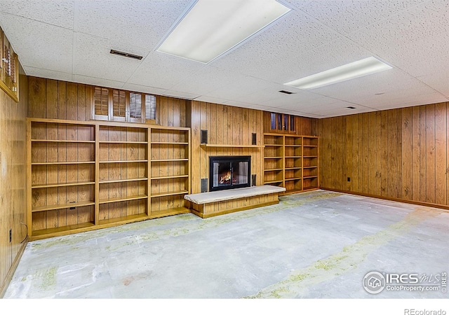 unfurnished living room featuring a drop ceiling, wooden walls, and concrete floors