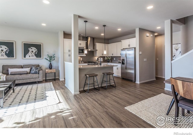 kitchen featuring pendant lighting, wall chimney range hood, white cabinetry, stainless steel appliances, and a center island with sink