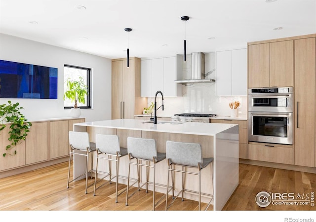 kitchen featuring wall chimney range hood, hanging light fixtures, stainless steel appliances, an island with sink, and white cabinets