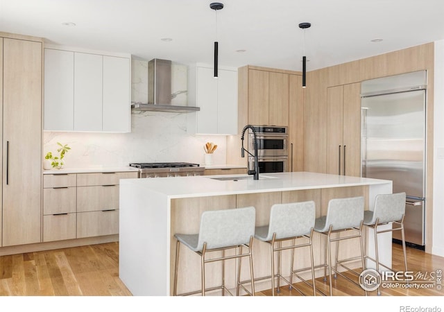 kitchen with appliances with stainless steel finishes, white cabinets, hanging light fixtures, a kitchen island with sink, and wall chimney range hood