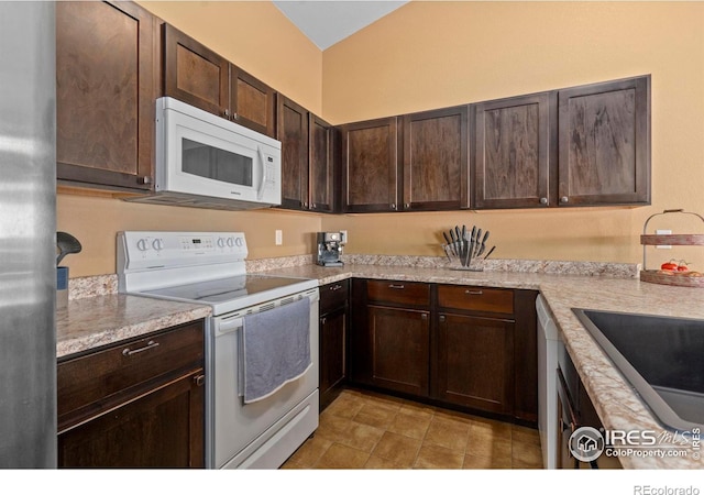 kitchen with lofted ceiling, dark brown cabinets, light stone counters, and white appliances