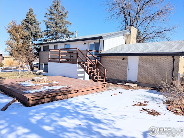 snow covered house with brick siding, stairs, a chimney, and a wooden deck