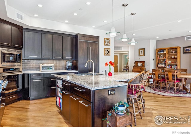 kitchen with a kitchen island with sink, dark brown cabinets, built in appliances, decorative backsplash, and decorative light fixtures