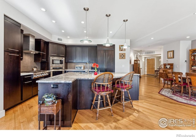 kitchen with decorative light fixtures, backsplash, built in appliances, light wood-type flooring, and wall chimney exhaust hood