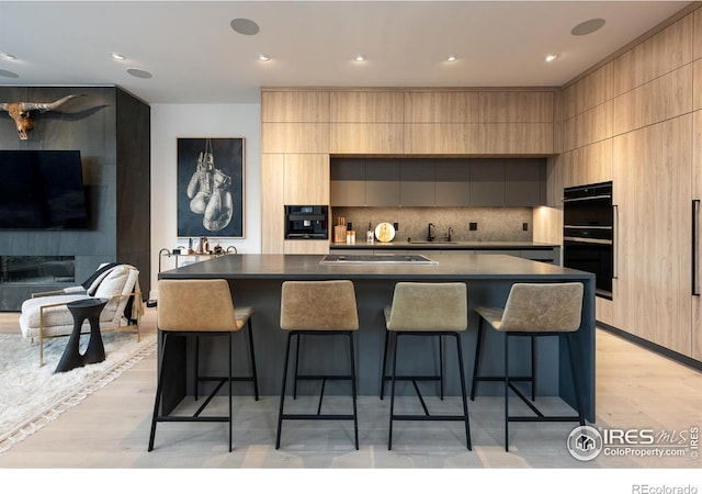 kitchen featuring black double oven, an island with sink, light brown cabinetry, and a kitchen breakfast bar