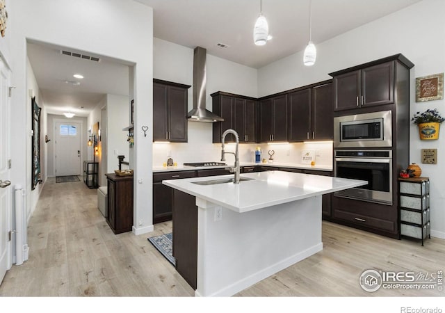 kitchen featuring appliances with stainless steel finishes, a kitchen island with sink, wall chimney range hood, and dark brown cabinets