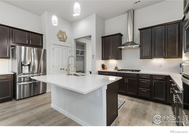 kitchen with sink, light wood-type flooring, appliances with stainless steel finishes, a kitchen island with sink, and wall chimney range hood