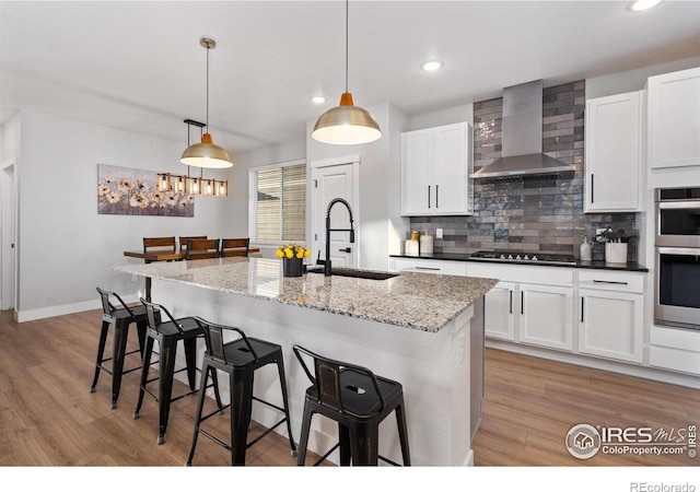 kitchen with appliances with stainless steel finishes, decorative light fixtures, sink, a kitchen island with sink, and wall chimney exhaust hood