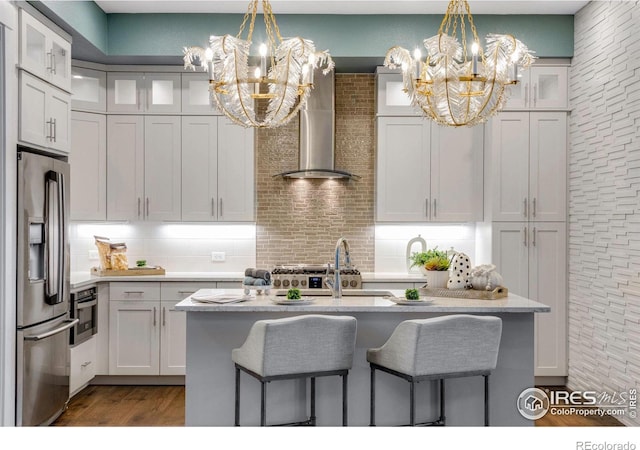 kitchen with an inviting chandelier, wall chimney range hood, a center island with sink, and appliances with stainless steel finishes