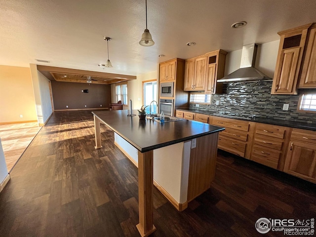 kitchen with appliances with stainless steel finishes, hanging light fixtures, tasteful backsplash, an island with sink, and wall chimney exhaust hood