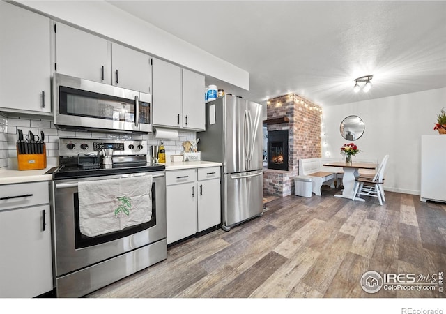 kitchen with hardwood / wood-style floors, white cabinetry, backsplash, stainless steel appliances, and a brick fireplace