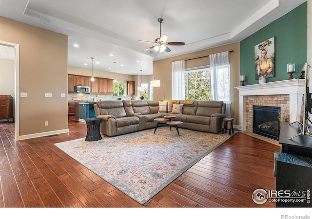 living room with a raised ceiling, a brick fireplace, dark wood-type flooring, and ceiling fan