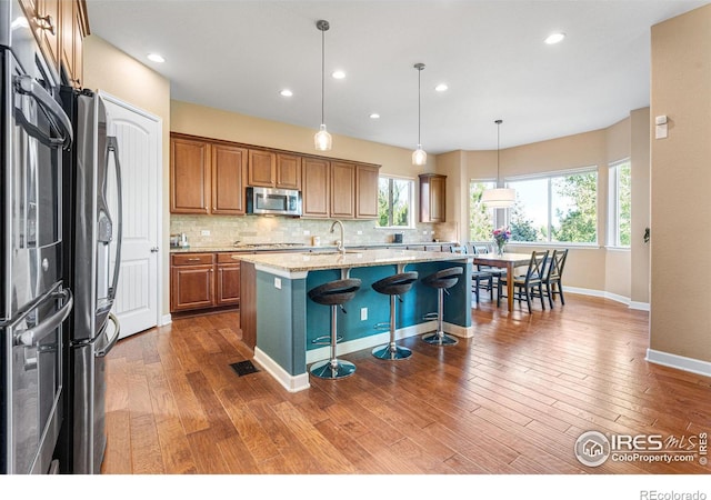 kitchen featuring tasteful backsplash, decorative light fixtures, a center island with sink, stainless steel appliances, and light stone countertops