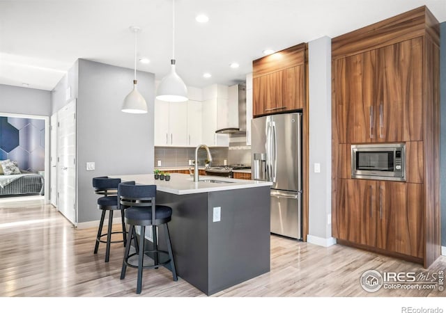 kitchen with wall chimney range hood, white cabinetry, a kitchen island with sink, stainless steel appliances, and decorative light fixtures