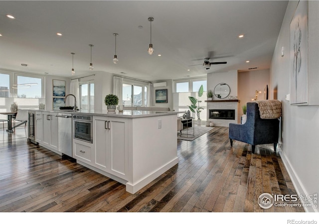 kitchen featuring appliances with stainless steel finishes, white cabinetry, an island with sink, a large fireplace, and dark hardwood / wood-style flooring