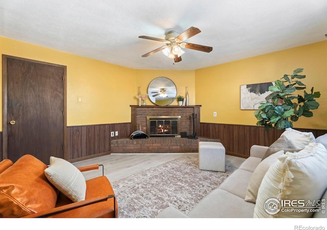 living room with ceiling fan, wooden walls, a textured ceiling, a brick fireplace, and light wood-type flooring