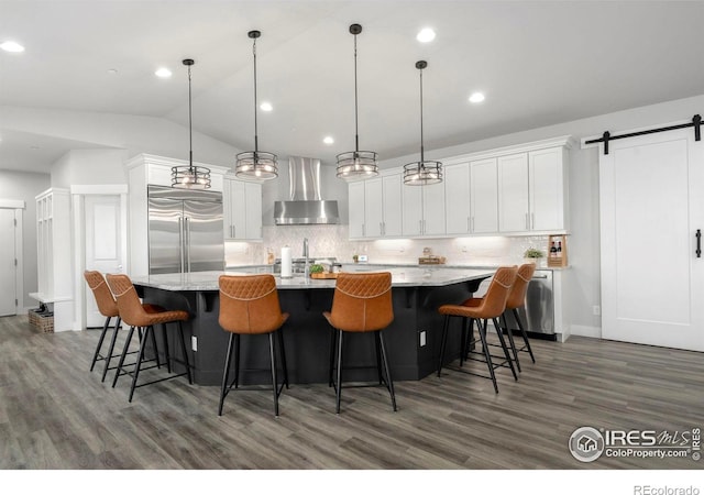 kitchen with a large island, built in refrigerator, decorative light fixtures, a barn door, and wall chimney exhaust hood