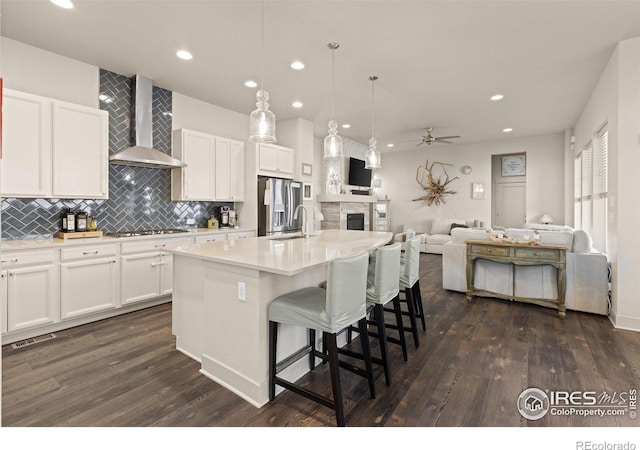 kitchen with white cabinetry, wall chimney exhaust hood, a center island with sink, and stainless steel refrigerator with ice dispenser