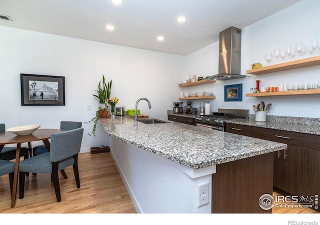 kitchen with sink, stainless steel range, light stone counters, dark brown cabinets, and wall chimney range hood