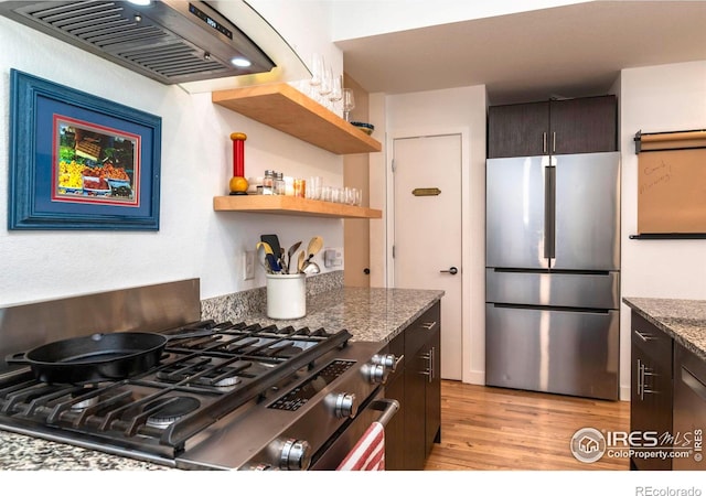 kitchen with appliances with stainless steel finishes, dark stone countertops, dark brown cabinets, exhaust hood, and light wood-type flooring