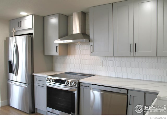 kitchen featuring wall chimney exhaust hood, light stone counters, tasteful backsplash, appliances with stainless steel finishes, and gray cabinets