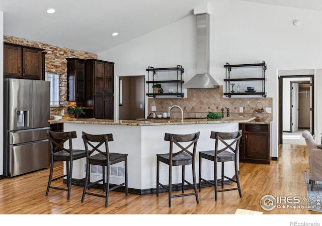 kitchen with stainless steel fridge, a kitchen island with sink, dark brown cabinetry, a kitchen bar, and exhaust hood