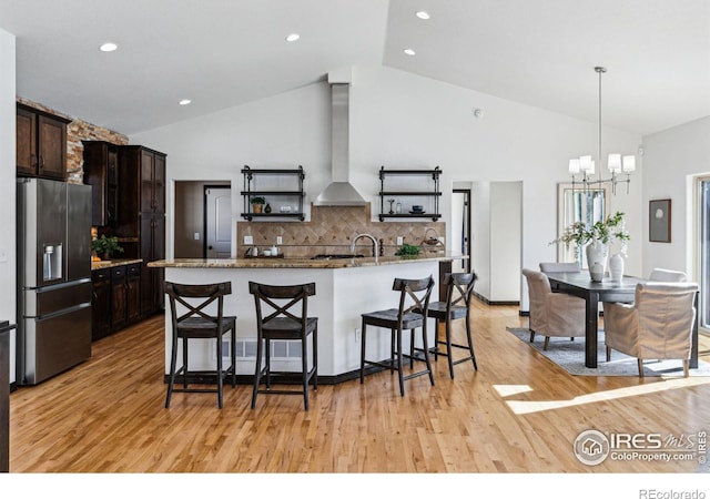 kitchen featuring decorative light fixtures, range hood, dark brown cabinetry, stainless steel refrigerator with ice dispenser, and a center island with sink