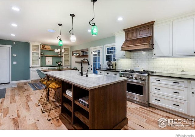 kitchen with white cabinetry, sink, high end stainless steel range oven, hanging light fixtures, and a center island with sink