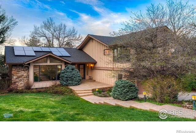 view of front of property with a shingled roof, stone siding, roof mounted solar panels, board and batten siding, and a front yard