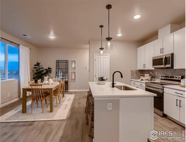 kitchen with appliances with stainless steel finishes, white cabinetry, sink, hanging light fixtures, and a center island with sink