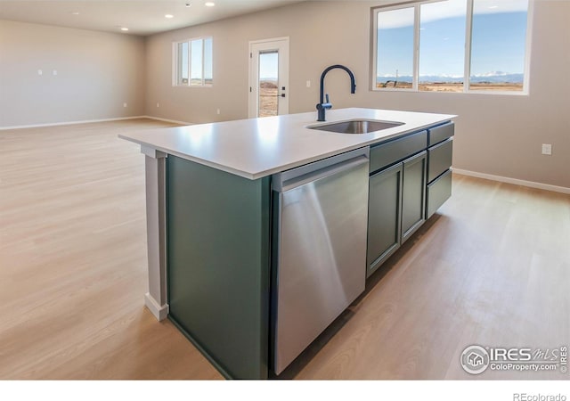 kitchen with light wood finished floors, a center island with sink, a sink, stainless steel dishwasher, and light countertops