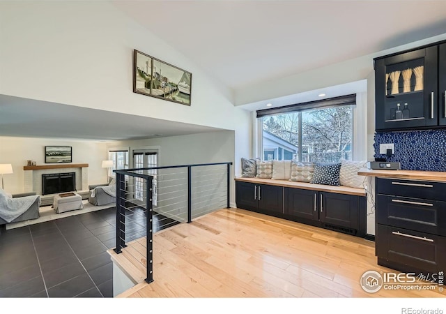 interior space with tasteful backsplash, a fireplace with raised hearth, glass insert cabinets, light wood-style flooring, and dark cabinets