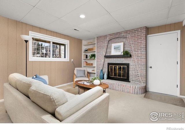 carpeted living room with built in shelves, a fireplace, a drop ceiling, and wood walls