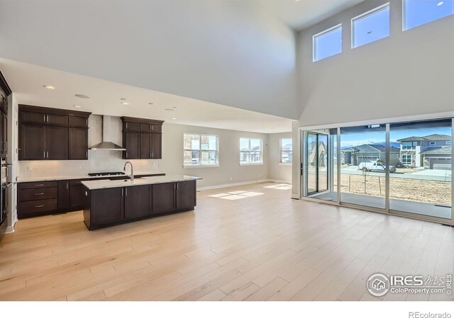 kitchen featuring dark brown cabinetry, open floor plan, light countertops, wall chimney range hood, and an island with sink