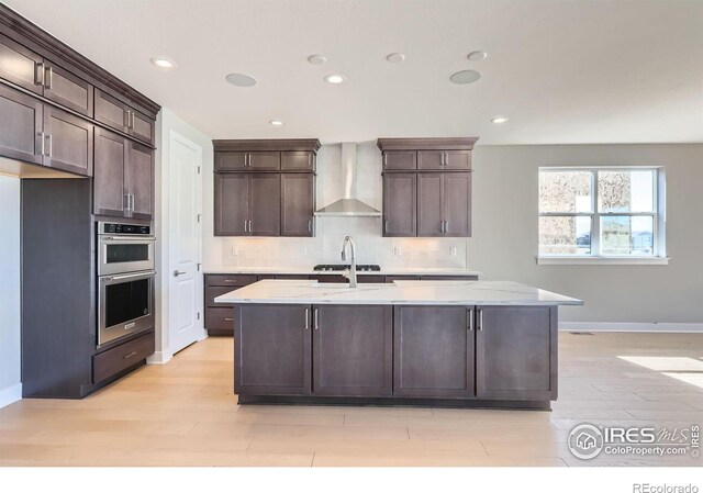 kitchen featuring backsplash, light wood-style flooring, double oven, dark brown cabinetry, and wall chimney range hood