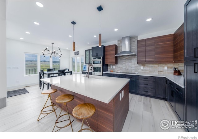 kitchen featuring a breakfast bar, tasteful backsplash, a kitchen island with sink, black electric stovetop, and wall chimney exhaust hood